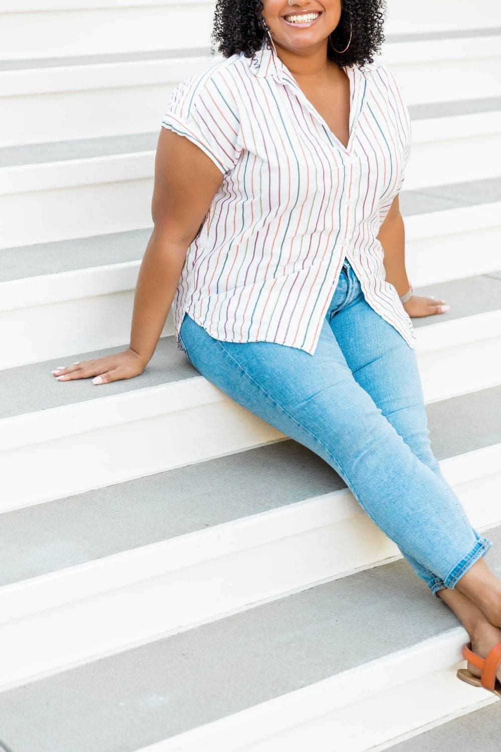 An image of an apple shaped black woman sitting on steps wearing a striped top and jeans illustrating the best jeans for an apple shape